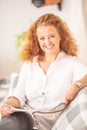 Woman with curly hair reads a magazine, sitting on a sofa in a living room, smiling at the camera Royalty Free Stock Photo