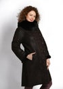 Woman with curly red hair in brown sheepskin coat with fur collar and traditional emboss pattern stands sideways posing