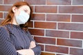 A woman in dark clothing and medical mask stands near a red brick wall, copy space for text