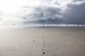 Woman in distance standing against rough sea on very long beach under dramatic cloudy sky with straight track of footprints