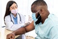 Woman doctor, wearing a surgical mask, is administering a vaccination treatment to an African American man patient