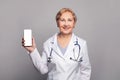 Woman doctor in white coat smiling while showing screen of his phone, using medical app, standing isolated on gray background Royalty Free Stock Photo