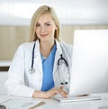 Woman-doctor at work while sitting at the desk in hospital or clinic. Blonde cheerful physician using desktop computer Royalty Free Stock Photo