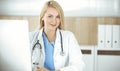 Woman-doctor at work while sitting at the desk in hospital or clinic. Blonde cheerful physician using desktop computer Royalty Free Stock Photo