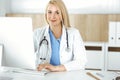 Woman-doctor at work while sitting at the desk in hospital or clinic. Blonde cheerful physician using desktop computer Royalty Free Stock Photo