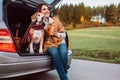 Woman and dog with shawls sits together in car trunk on autumn