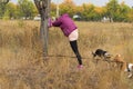 Woman doing stretching exercise near wild pear tree while walking outdoors with pair of dogs in autumnal park