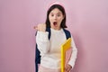 Woman with down syndrome wearing student backpack and holding books surprised pointing with hand finger to the side, open mouth Royalty Free Stock Photo