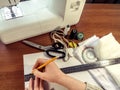 Woman draws a sewing pattern with a pencil and a ruler on white paper against a sewing machine and accessories lying on a brown