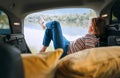 Woman dressed in warm calm colors clothes and jeans lying in the cozy car trunk and enjoying the mountain lake view. Warm early