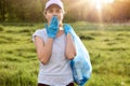 Woman dresses baseball cap and casual t shirt, holding garbage bag, covering mouth with palm wears blue latex glove, being in