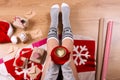 Woman drinking cappuccino coffee and sitting on the wooden floor. Close-up of female legs in warm socks with a deer with christmas