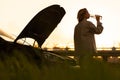 Woman drinking water, waiting for assistance near her car broken down with open bonnet at sunset Royalty Free Stock Photo