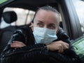 Woman driver in a medical mask sits in the car in front of and looks out the window. Quarantined security. Royalty Free Stock Photo