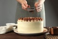 Woman dusting delicious tiramisu cake with cocoa powder at wooden table, closeup