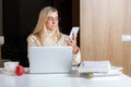 Woman in earphones using cellphone while working with laptop at home kitchen Royalty Free Stock Photo