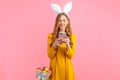 Woman in the ears of an Easter Bunny, holding a basket of Easter eggs, with a mobile phone in her hands on an pink background