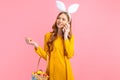 Woman in the ears of an Easter Bunny holds a basket of colorful Easter eggs and talks on a mobile phone, on an pink background