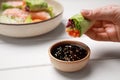 Woman eating delicious spring roll wrapped in rice paper with sauce at white wooden table, closeup. Space for text Royalty Free Stock Photo