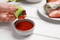 Woman eating delicious spring roll wrapped in rice paper with sauce at white wooden table, closeup Royalty Free Stock Photo