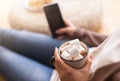 Woman enjoying cocoa with marshmallow and using cellphone