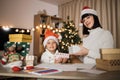 Woman exchanging sharing gifts with her daughter in santa hats while sitting near decorated tree Royalty Free Stock Photo