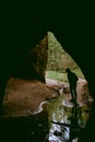 A woman exploring a dark cave with flowing water and forest in the background. The Devil`s Oven in Liepa, Latvia