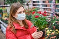 A woman in a face mask chooses red roses grown in the greenhouse of a flower shop. The sales of potted plants for a hobby during Royalty Free Stock Photo