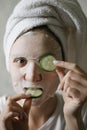 A woman in a face mask is having fun with round cucumbers, looking at the camera and biting a cucumber. Natural