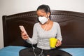 Woman with face mask and sitting on the bed talking on her cell phone at home in her bedroom Royalty Free Stock Photo