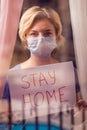 Woman with face medical mask staying in flat in front of window holding a sheet of paper with title stay home. People and