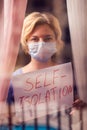 Woman with face medical mask staying in flat in front of window holding a sheet of paper with title self-isolation. People and