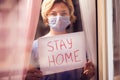 Woman with face medical mask staying in flat in front of window holding a sheet of paper with title stay home. People and