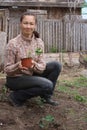 Woman farmer sitting next to strawberry garden bed and holding pot with strawberry seedling ready to plant it to soil