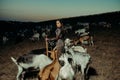 Woman farmer stands with pitchfork among goats on pasture in the night