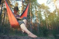 Woman in fedora hat reading book while sitting in hammock in pine forest at sunset. Mindfulness, technology break, wellbeing, Royalty Free Stock Photo