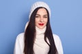 Woman feeling cold, wearing white cap, sweater and scarf, looking directly directly at camera with charming smile and red lips, Royalty Free Stock Photo