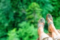 Woman feet in sandals dangling from steep edge of mountain rock on green forest trees background