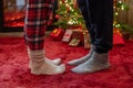 Woman feet standing in tip toe in winter socks on male lags on a fluffy red blanket near a Christmas tree with gifts. Concept. Royalty Free Stock Photo