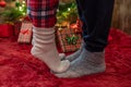 Woman feet standing in tip toe in winter socks on male lags on a fluffy red blanket near a Christmas tree with gifts. Concept. Royalty Free Stock Photo