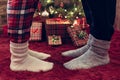 Woman feet standing in tip toe in winter socks on male lags on a fluffy red blanket near a Christmas tree with gifts. Concept. Royalty Free Stock Photo