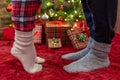 Woman feet standing in tip toe in winter socks near male lags on a fluffy red blanket near a Christmas tree with gifts. Concept. Royalty Free Stock Photo