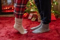 Woman feet standing in tip toe in winter socks near male lags on a fluffy red blanket near a Christmas tree with gifts. Concept. Royalty Free Stock Photo