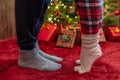 Woman feet standing in tip toe in winter socks near male lags on a fluffy red blanket near a Christmas tree with gifts. Concept. Royalty Free Stock Photo