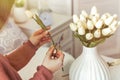 Woman florist cutting stem of tulips flowers with scissors and putting in vase on coffee table. Composing bouque.