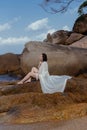 Woman in a flowing white dress enjoys a peaceful moment on coastal rocks, the sea softly lapping near her Royalty Free Stock Photo