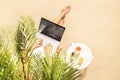 Woman freelancer with laptop sitting under palm tree branches. Sunscreen, sunglasses, orange juice on the table of sandy beach Royalty Free Stock Photo