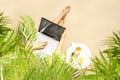 Woman freelancer with laptop sitting under palm tree branches. Sunscreen, sunglasses, orange juice on the table of sandy beach Royalty Free Stock Photo