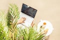 Woman freelancer with laptop sitting under palm tree branches. Sunscreen, sunglasses, orange juice on the table of sandy beach Royalty Free Stock Photo