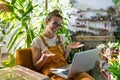 Woman gardener sitting on chair in green house, using laptop after work, smiling and speaking on video call.Home gardening, small